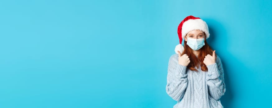 Christmas, quarantine and covid-19 concept. Cute teen redhead girl in santa hat and sweater, wearing face mask from coronavirus, showing thumbs up, standing over blue background.