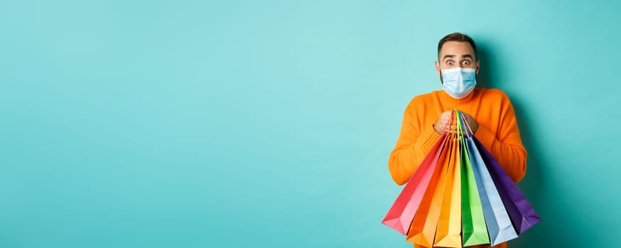 Covid-19, pandemic and lifestyle concept. Excited man in face mask showing shopping bags and rejoicing from discounts, standing over turquoise background.