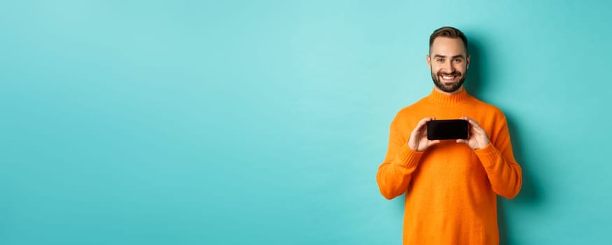 Handsome bearded adult man showing mobile screen, holding smartphone horizontally, concept of applications and online shopping.