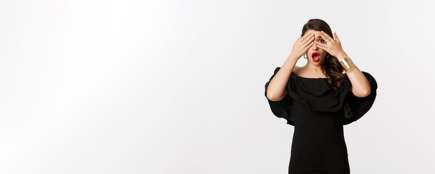 Fashion and beauty. Image of shocked glamour woman in black dress, covering eyes but peeking through fingers startled, standing over white background.