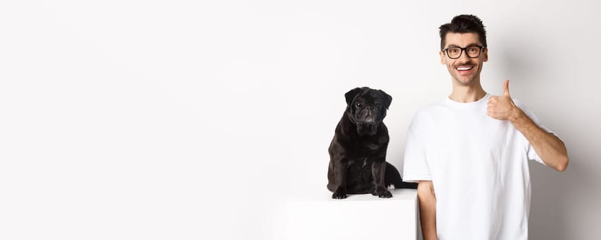 Image of dog owner and cute black pug looking at camera, man showing thumb-up in approval, recommending something, standing over white background.