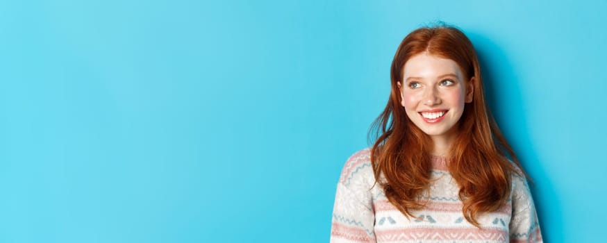 Close-up of beautiful redhead female model in winter sweater, smiling and looking left at copy space, standing over blue background.