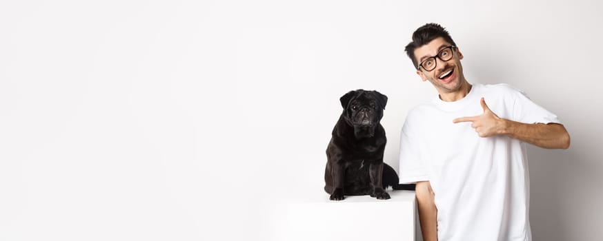 Happy young man showing his cute dog, pointing finger at black pug and smiling, standing over white background.