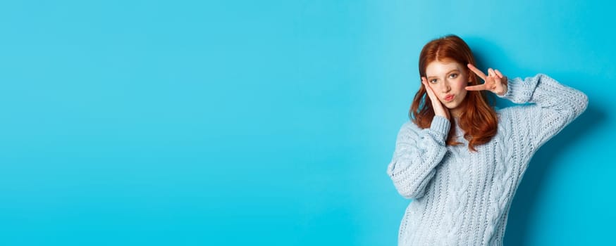 Modern teen girl with red hair, showing peace sign and posing in sweater against blue background.