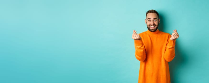 Smiling handsome man showing hearts and looking happy, lucky gesture, standing in orange sweater over light blue background.