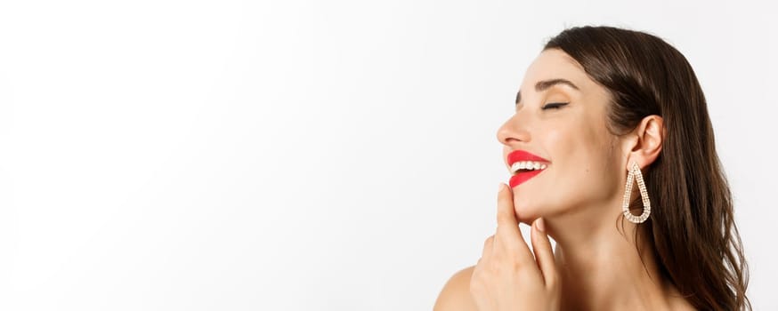 Fashion and beauty concept. Headshot of sensual brunette woman with earrings and red lipstick, smiling tempted and touching lip, standing over white background.