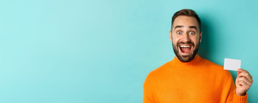 Close-up of excited caucasian man showing his credit card, smiling and staring amazed, standing in orange sweater against turquoise background.