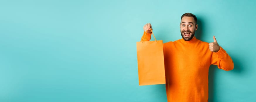 Happy man holding orange shopping bag and rejoicing from discount and celebrating, showing thumbs up and recommending, standing over blue background.