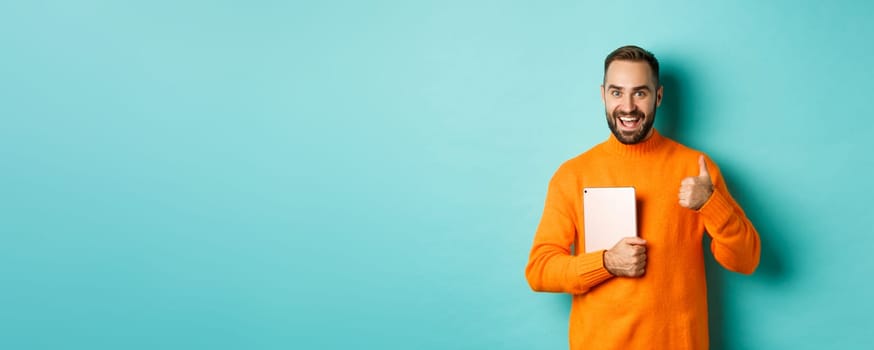 Work from home, technology concept. Handsome man holding laptop, showing thumb up, approve and like something, standing over turquoise background.