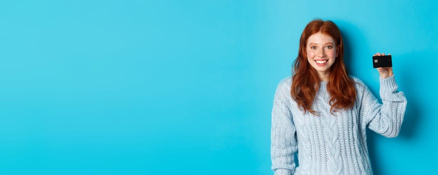 Cute redhead girl in sweater showing credit card, smiling at camera, standing over blue background.