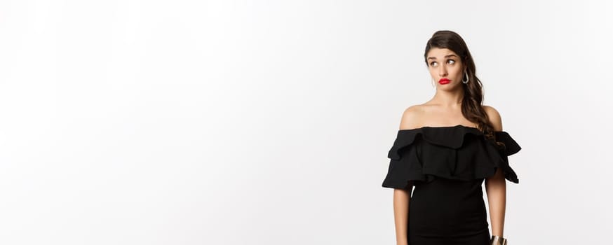 Fashion and beauty. Confused attractive woman in black dress, looking away with puzzled and shocked face, standing over white background.