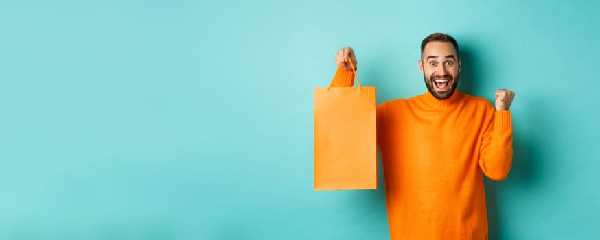 Happy man holding orange shopping bag and rejoicing, got discount and celebrating, standing over turquoise background.