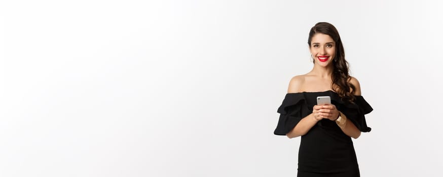 Online shopping concept. Attractive young woman in black dress, reading text message, using mobile phone and smiling, standing over white background.