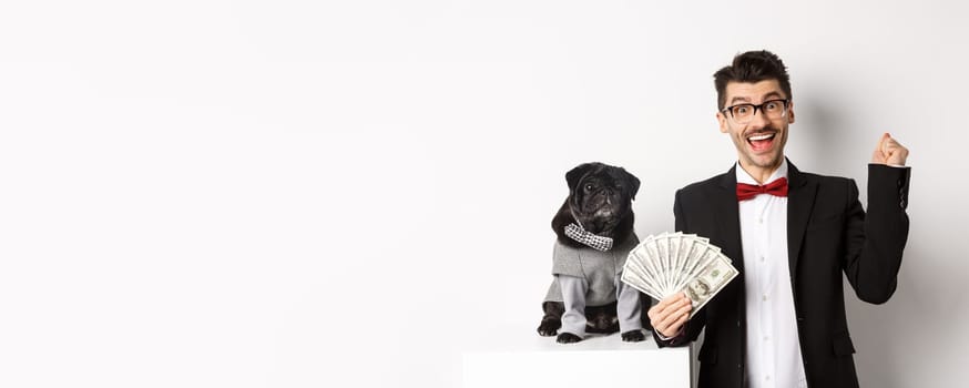 Happy young man in suit earn money with his dog. Guy rejoicing, holding dollars, black pug in costume staring at camera, white background.