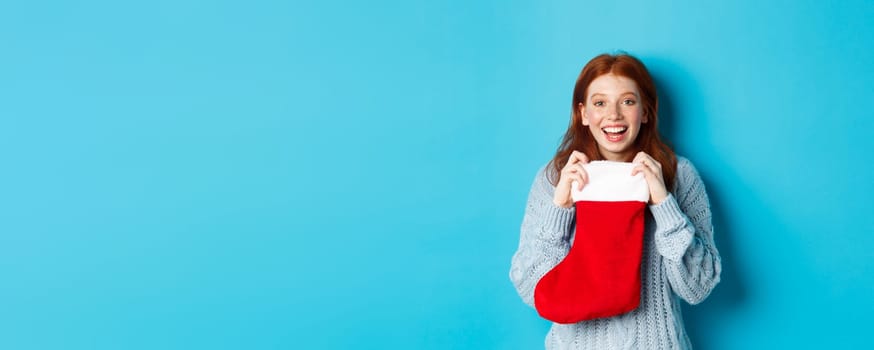 Winter holidays and gifts concept. Happy teenage redhead girl receiving xmas gift, open christmas stocking and smiling amazed, standing over blue background.