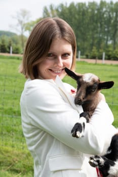 Young woman plays with goat kids, feeding them, sun shining over farm in background, High quality photo