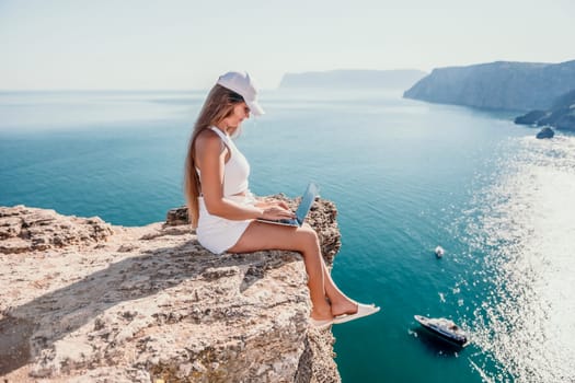 Successful business woman in yellow hat working on laptop by the sea. Pretty lady typing on computer at summer day outdoors. Freelance, travel and holidays concept.