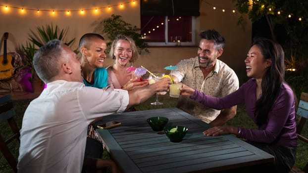 Diverse group of friends laughing and having fun during night garden party drinking cocktails. Evening party.