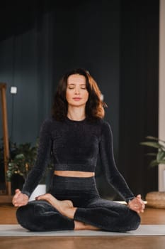 A woman in a black tracksuit sits on a yoga mat in a lotus position in the gym.