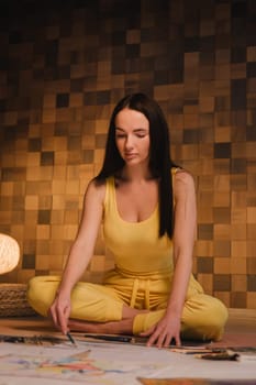 A girl in yellow clothes sitting in the Lotus position draws on paper with evening lighting at home.