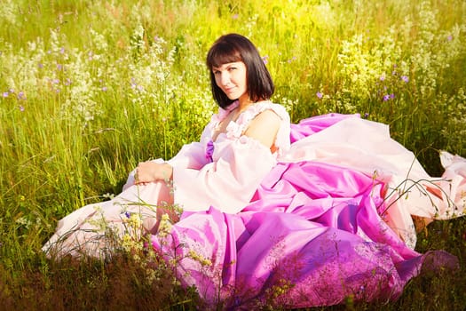 Beautiful girl in lush pink ball gown in green field during blooming of flowers and blue sky on background. Model posing on nature landscape as princess from fary tale