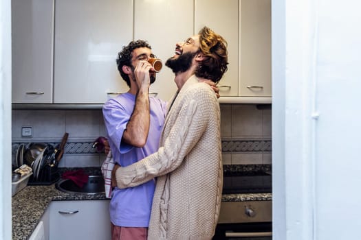 gay male couple hugging and laughing happy while having morning coffee in the kitchen, concept of home lifestyle and love between people of the same sex, copy space for text
