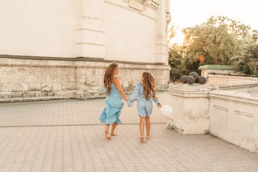 Daughter mother run holding hands. In blue dresses with flowing long hair, they hold balloons in their hands against the backdrop of a sunset and a white building