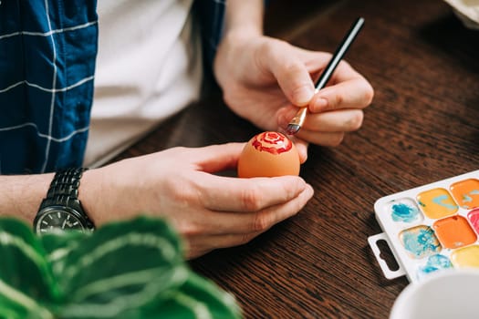 Easter day. Cropped hands of Adult man painting eggs on wooden background. Sitting in a kitchen with crayons. Preparing for Easter, creative homemade decoration.