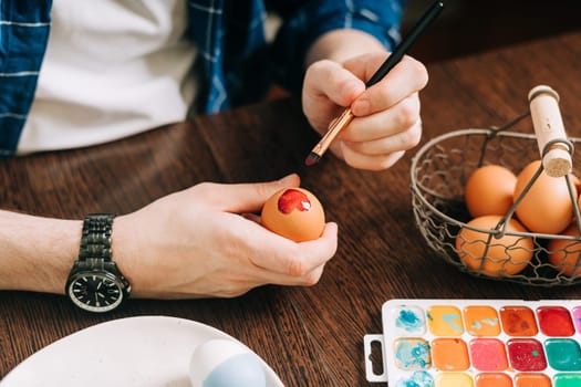 Easter day. Cropped hands of Adult man painting eggs on wooden background. Sitting in a kitchen with crayons. Preparing for Easter, creative homemade decoration.