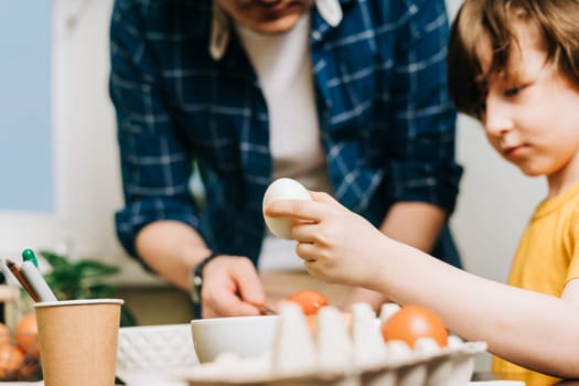 Easter day. Male Father and son painting eggs on wooden background. Family sitting in a kitchen. Preparing for Easter, creative homemade decoration. Child kid boy having fun and painting easter eggs