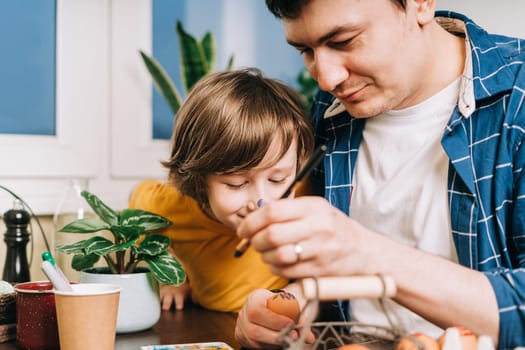 Easter day. Male Father and son painting eggs on wooden background. Family sitting in a kitchen. Preparing for Easter, creative homemade decoration. Child kid boy having fun and painting easter eggs