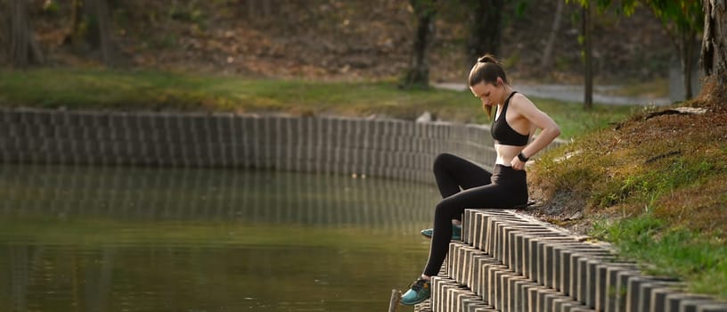 Fitness woman sitting near lake resting after successful jogging exercise outdoors. Horizontal photo banner for website header design.