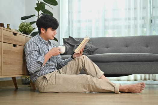 Full length of man in casual wear drinking coffee and enjoying reading interesting book in modern trendy cozy home interior.