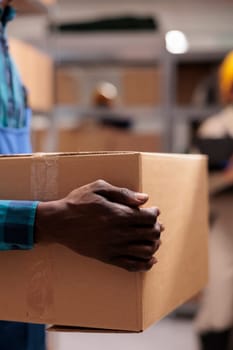 Industrial warehouse employee holding cardboard package ready for shipment. African american postal office worker arms carrying customer parcel in mail sorting department storage closeup