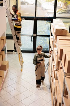 Supervisor scanning carton boxes using store scanner during storehouse inventory, typing products barcode on tablet computer. Employee in protective overall preparing customers orders in warehouse
