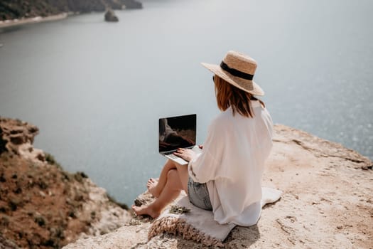 Successful business woman in yellow hat working on laptop by the sea. Pretty lady typing on computer at summer day outdoors. Freelance, travel and holidays concept.