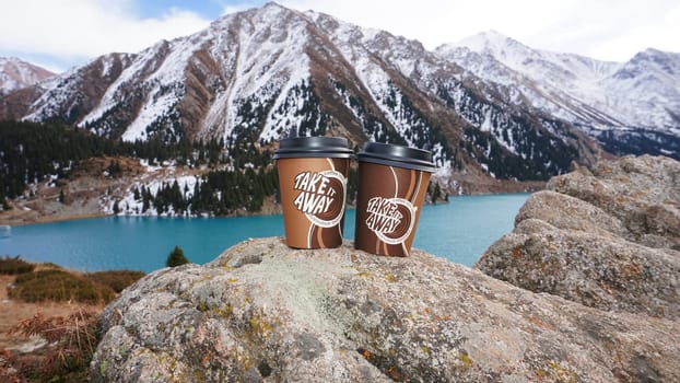 Paper cups with coffee on the background of a mountain lake. The color of the water is blue. A green forest grows on the hills. The high peaks are covered with snow. The cups are on a stone. Romance