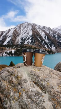 Paper cups with coffee on the background of a mountain lake. The color of the water is blue. A green forest grows on the hills. The high peaks are covered with snow. The cups are on a stone. Romance