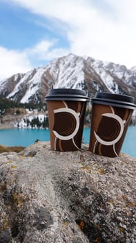 Paper cups with coffee on the background of a mountain lake. The color of the water is blue. A green forest grows on the hills. The high peaks are covered with snow. The cups are on a stone. Romance