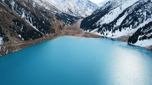 A lake in the mountains with turquoise blue water. Drone view of clear water, coniferous trees and snowy mountains. People walk along the shore, low bushes grow. Big Almaty lake. Kazakhstan