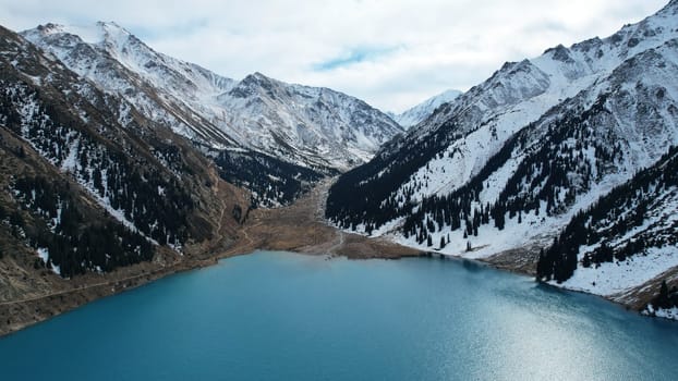 A lake in the mountains with turquoise blue water. Drone view of clear water, coniferous trees and snowy mountains. People walk along the shore, low bushes grow. Big Almaty lake. Kazakhstan