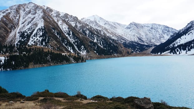 A mountain lake with blue water in winter. The sun's rays are reflected from the water. Green forest and grass grow on the hills. There are stones lying. The high peaks are covered with snow. Almaty