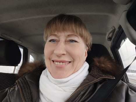 Portrait of adult cute funny blonde woman in warm clothes taking a selfie inside the car and driving it. The concept of pleasant trip in spring, autumn, winter