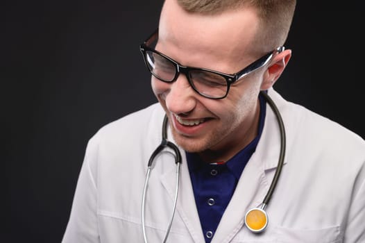 A young caucasian doctor in a white coat laughs and has fun. Portrait of a laughing doctor on a black background. Cheerful male doctor. Positive medical professional.