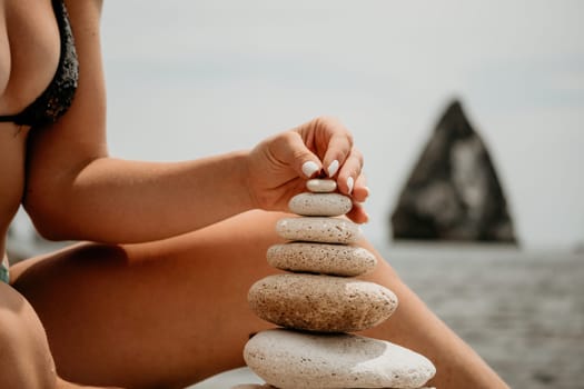 Woman bilds stones pyramid on the seashore on a sunny day on the blue sea background. Happy holidays. Pebble beach, calm sea, travel destination. Concept of happy vacation on the sea, meditation, spa