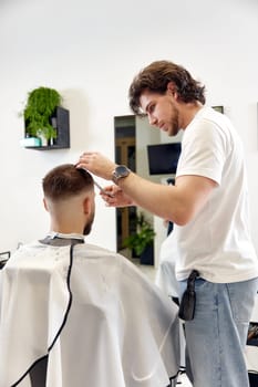 hairdresser does haircut for caucasian bearded man using comb and grooming scissors in barber shop.