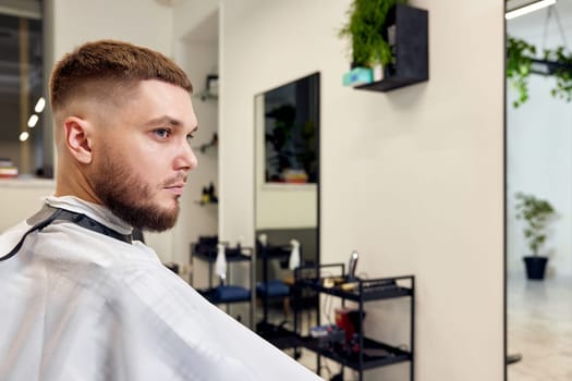 Handsome young man looks at his reflection in the mirror while sitting in chair in barbershop