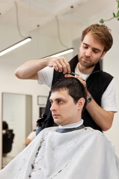 Professional hairdresser does haircut for caucasian client man at barber shop.