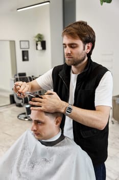 Professional hairdresser does haircut for caucasian bearded man using comb and scissors at barber shop.