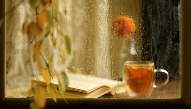 a cup of fragrant tea with an open book on a wooden windowsill against the background of an autumn rainy window